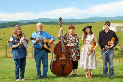 The members of the Whitetop Mountain Band hold their instruments and stand in a green field, mountain ranges visible in the distance behind them. From left, a young white man holding a fiddle in a black button down shirt and jeans, a young white woman with long brown hair holding a fiddle and wearing a light pink dress, a middle aged white woman with short red-brown hair and holding a double bass, an older white man with white hair holding an acoustic guitar and wearing a blue button down shirt and jeans, and a middle aged white woman with medium length brown hair holding a banjo and wearing a denim shirt and denim pants.